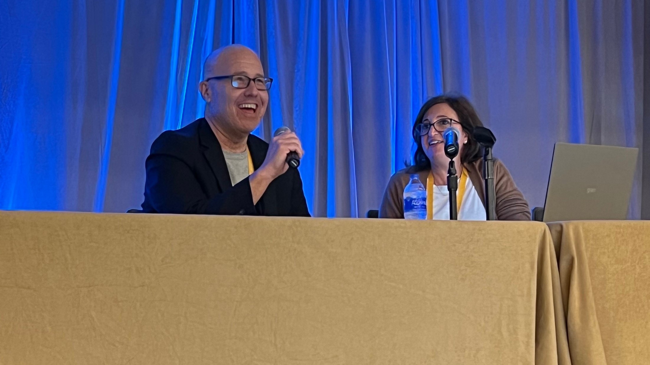 Bill Friedman y Ana Handshuh hablando juntos en una mesa con micrófonos en la conferencia RISE HEDIS 2022